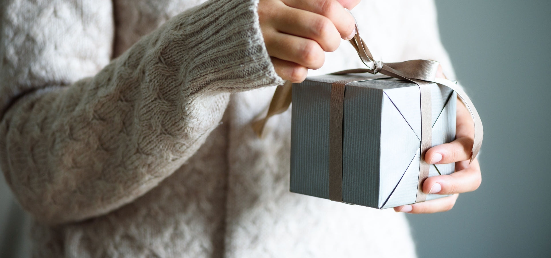 Person opening a gift box with a ribbon, wearing a beige sweater.
