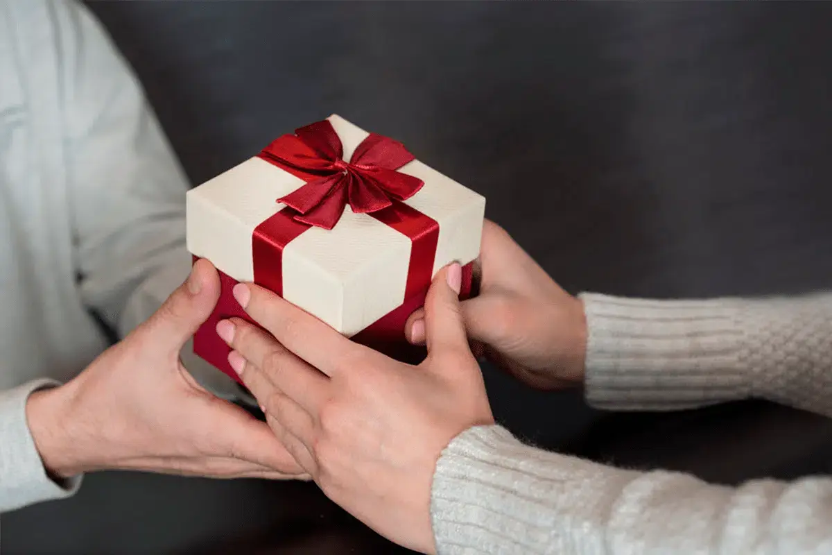 Two hands exchanging a hexagonal gift box with a red ribbon against a dark background